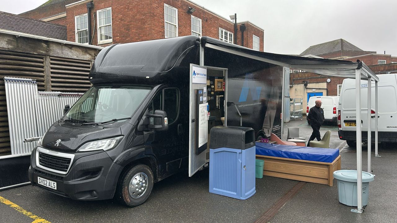 The ALS display vehicle parked on site with its awning extended and anti-ligature furniture set out, including a moulded bed, seating and waste bins