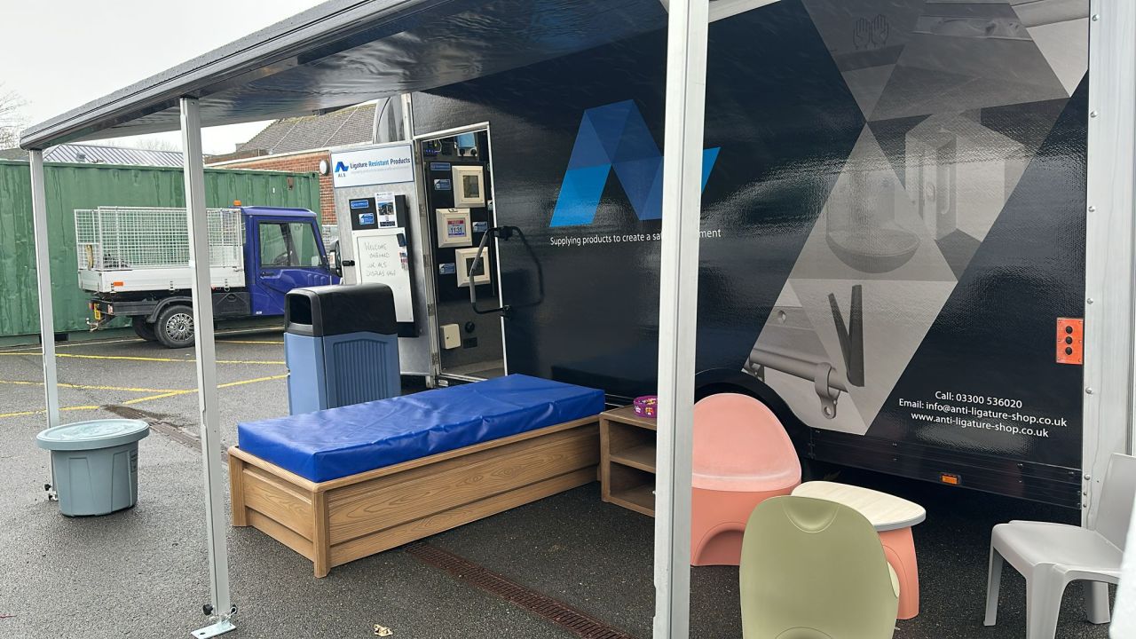 The ALS display vehicle set up on site with its awning out and anti-ligature furniture on display, including a moulded bed, nightstand and seating